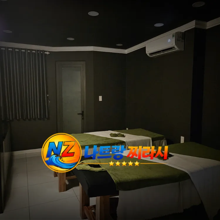 Dimly lit massage room with two treatment tables covered in green and white linens, dark walls, and an air conditioner mounted high on the wall; watermark logo at bottom.
