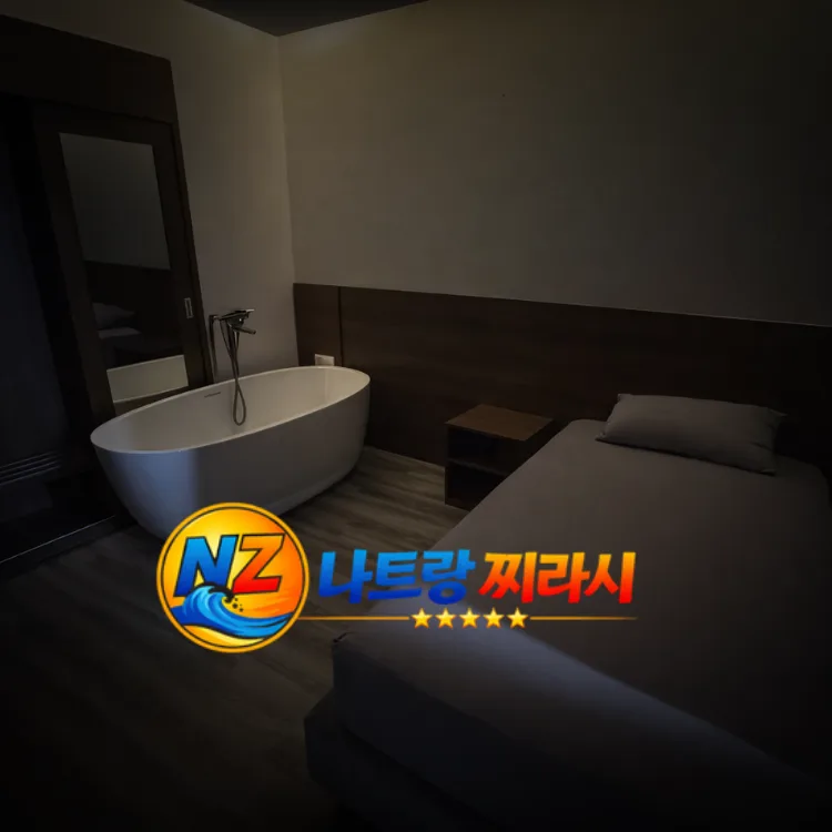 Dimly lit hotel room with a white freestanding bathtub beside the bed and a dark wood headboard. The logo overlays the lower left corner.