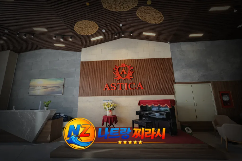 Lobby interior with a wooden wall displaying the red “ASTICA” logo, a black piano with a red cover, and a vase of flowers nearby.