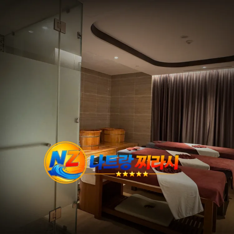 Spa treatment room with several massage beds, brown towels, and wooden buckets under warm lighting. A glass door and dark curtained wall are visible.