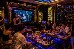 Woman sings karaoke into a microphone in a neon-lit VIP lounge while friends watch from a glossy table with drinks and snacks.